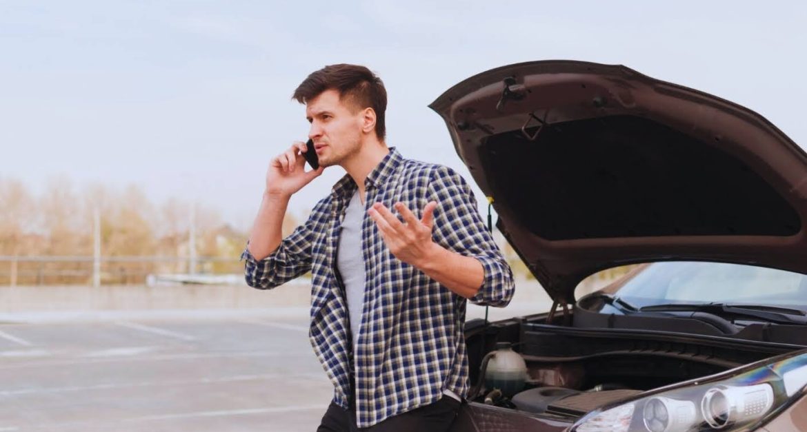Man on phone standing beside a car with the hood open, looking frustrated.