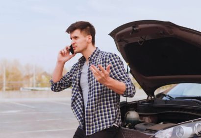 Man on phone standing beside a car with the hood open, looking frustrated.