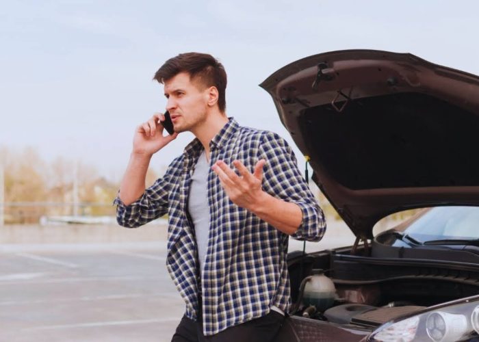 Man on phone standing beside a car with the hood open, looking frustrated.